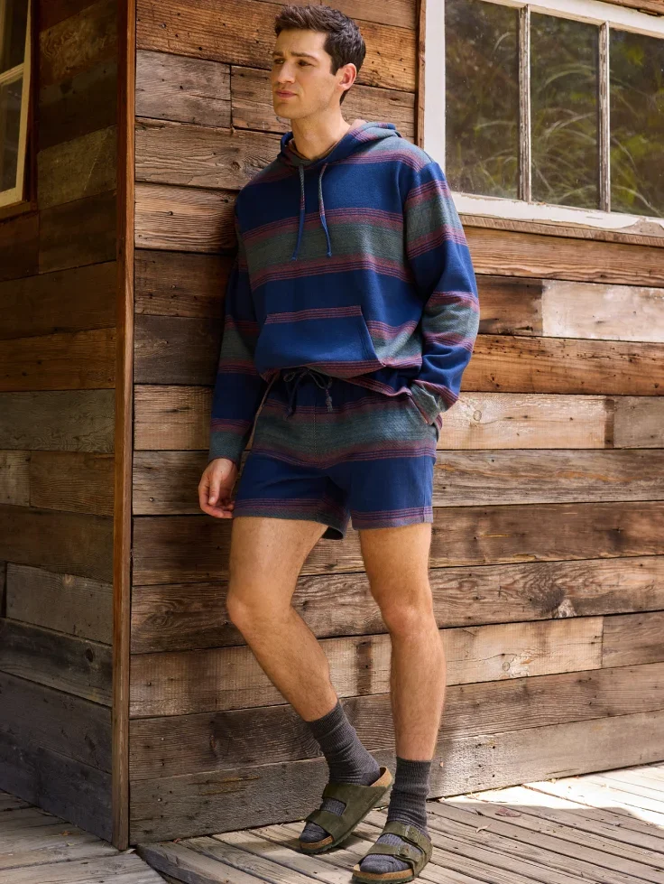 A man in a striped hoodie and shorts leans on a wooden wall
