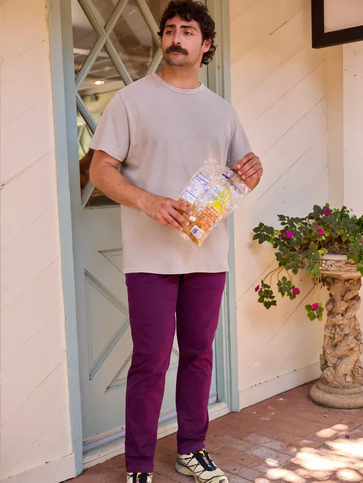 A man in a gray tee and plum pants holds a bag of snacks in a doorway
