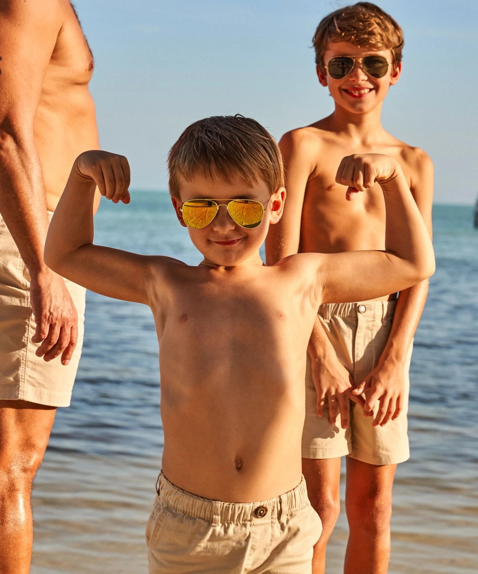 Two boys flexing their arms on the beach at sunset, one in yellow sunglasses