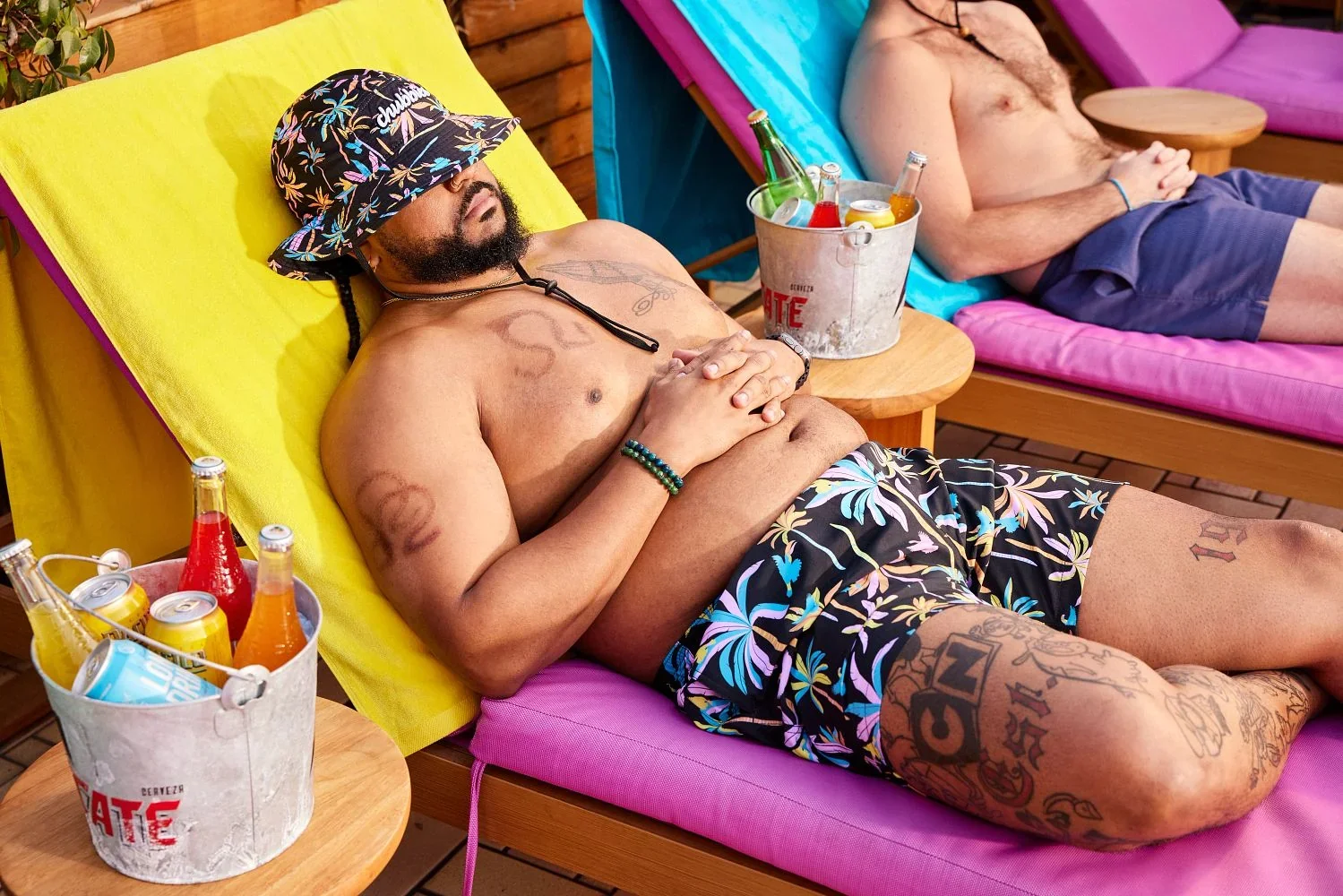 A man lounging by a pool in floral swim trunks and a bucket hat with a cooler of drinks