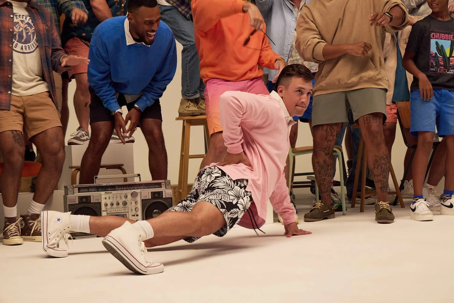 A crowd of friends in colorful casual clothes laughing around a boombox in a studio