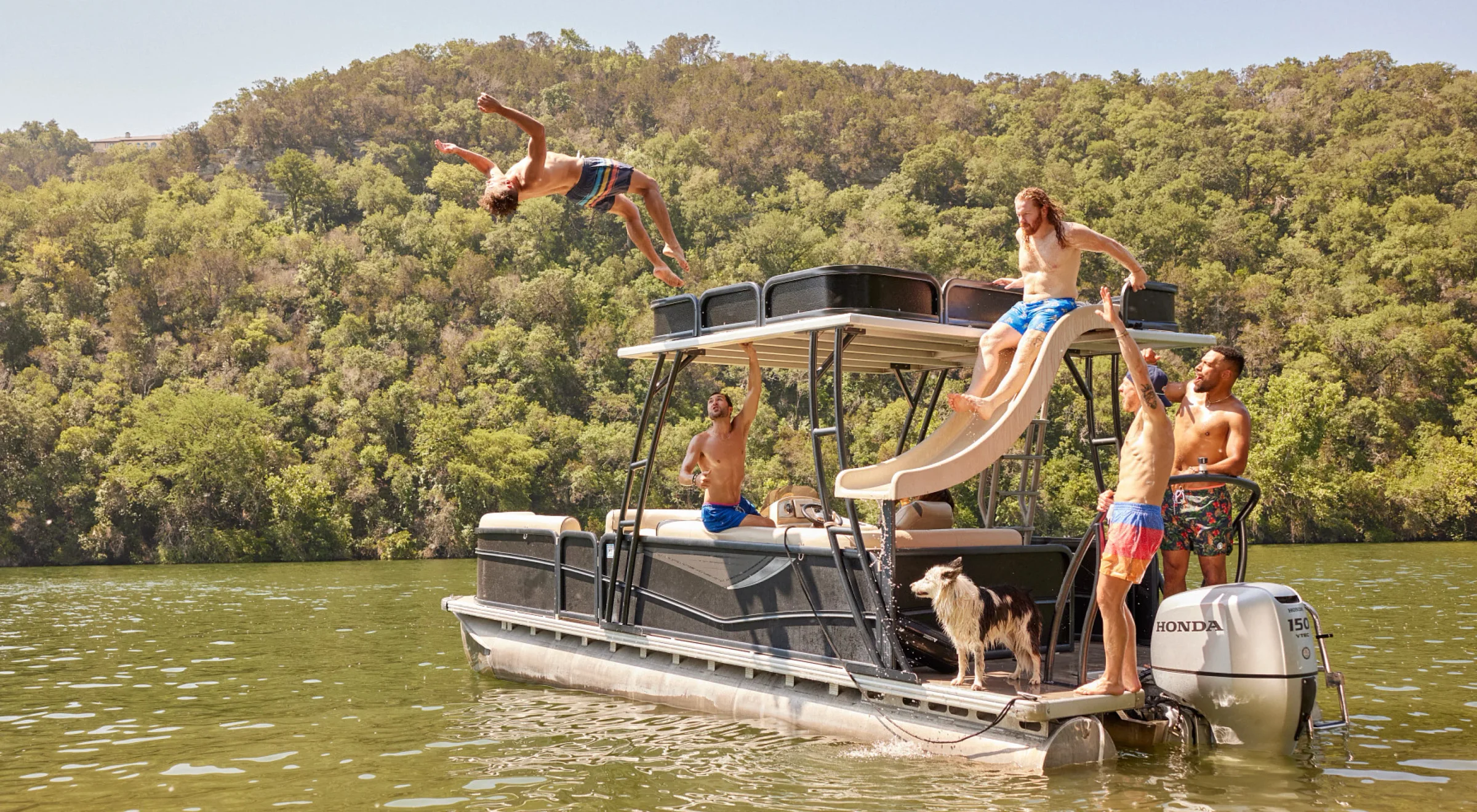 Friends jumping off the slide of a pontoon boat on a green lake