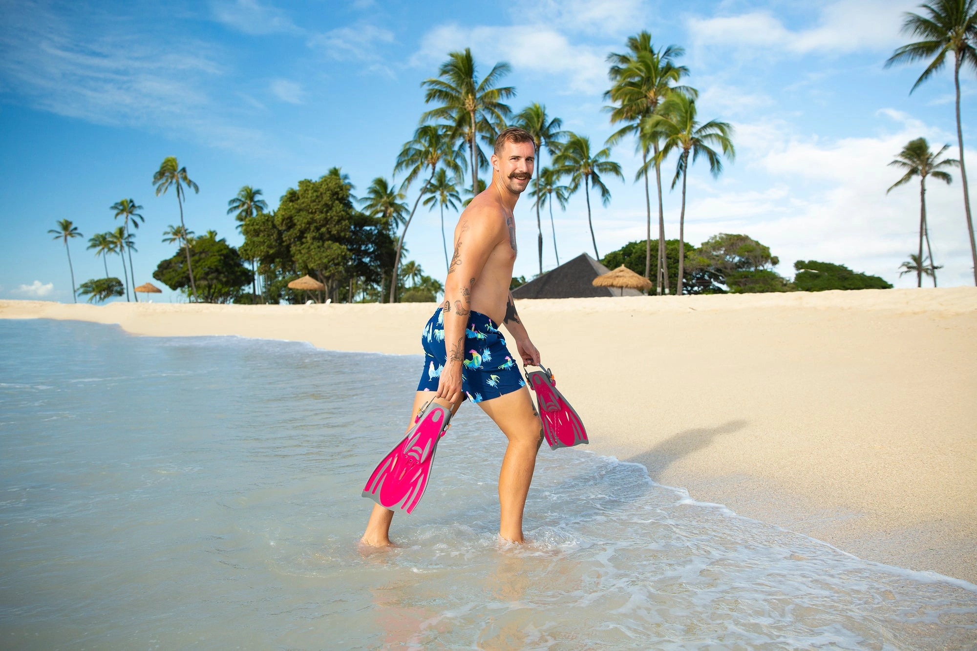 A man in flippers and swim trunks walks across a tropical beach framed by palm trees