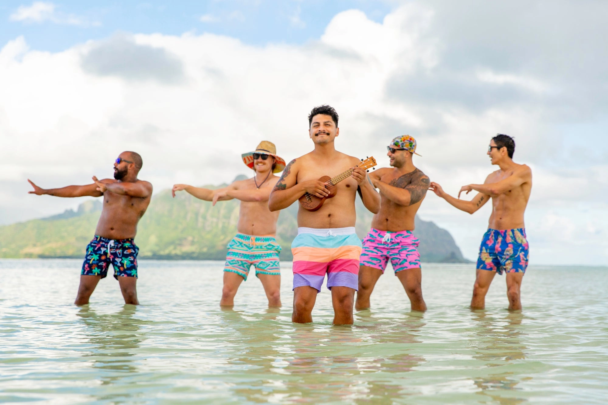 Six men standing in shallow turquoise water in bright swim trunks