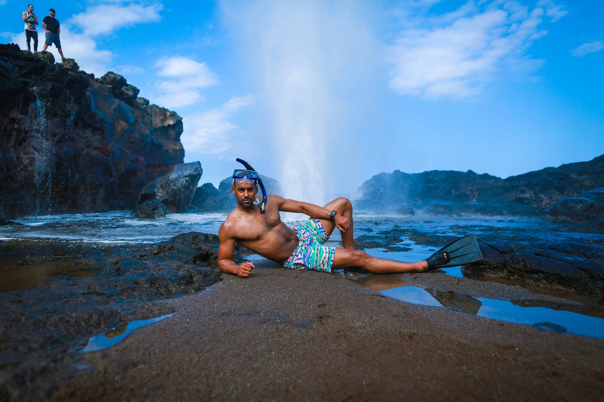 A man lounging on a rocky shore where waves burst behind him