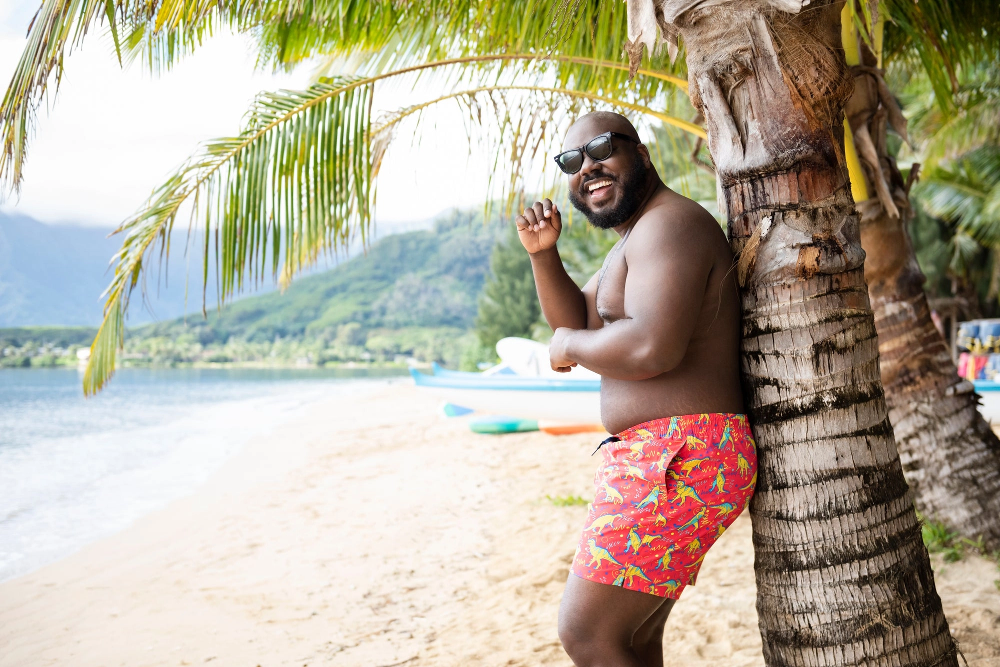 A smiling man in red swim trunks leaning against a palm tree
