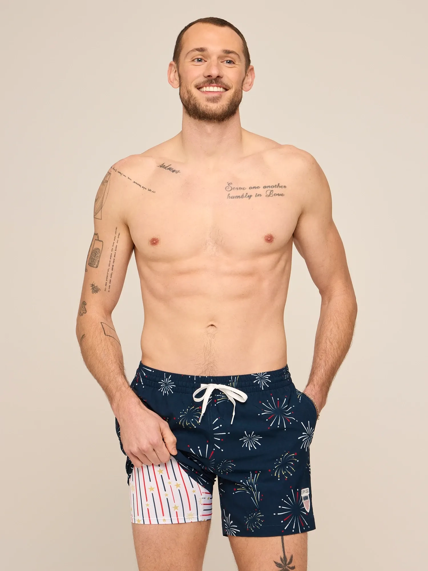A shirtless man in navy swim trunks with white stripes against a sand backdrop